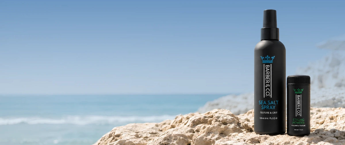 A black spray bottle and matching small container of Barbe & Co Sea Salt Spray standing on a rocky beach with the ocean and blue sky in the background.